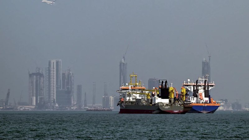 Two industrial vessels sit on hazy water with a distant city skyline and construction cranes visible in the background.