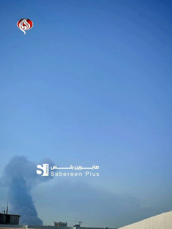 A plume of dark smoke rises against a clear blue sky above rooftop structures, with a news outlet's logo and Arabic text overlay visible.