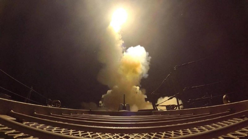 A missile launches skyward from the deck of a naval vessel at night, leaving a bright flame trail and billowing smoke against the dark sky.