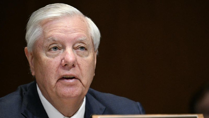 An older man with white hair, wearing a dark suit and white shirt, speaks against a dark background, his mouth slightly open.