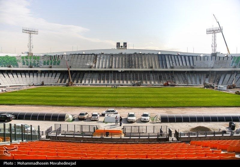 Maintenance crews work on the seating bowl and field of a large stadium with orange foreground seats under a partly cloudy sky.
