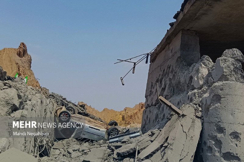 An overturned vehicle lies amid rubble and broken concrete beneath a damaged concrete structure under a clear sky.