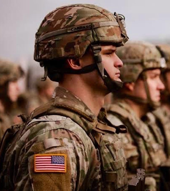 Soldiers in camouflage uniforms and helmets stand in formation, with the lead soldier's helmet displaying the name "Carrasco" and an American flag patch on the shoulder.
