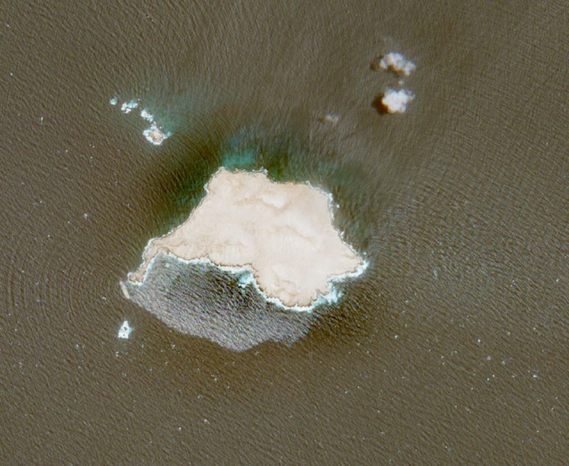 Aerial view of a tan landmass or salt formation surrounded by dark water, with smaller white deposits scattered nearby.