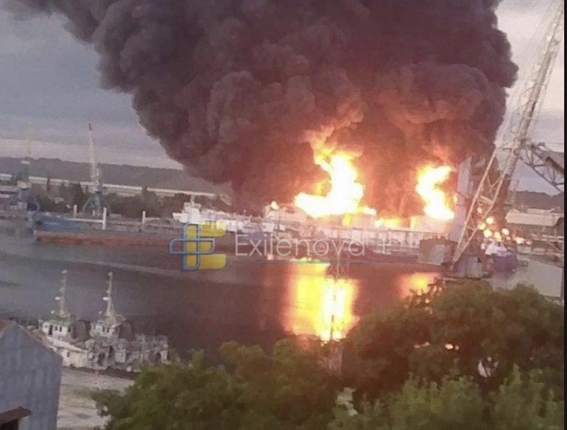 Large flames and thick black smoke rise from a docked ship at a harbor, with smaller vessels moored nearby and a crane visible on the right.