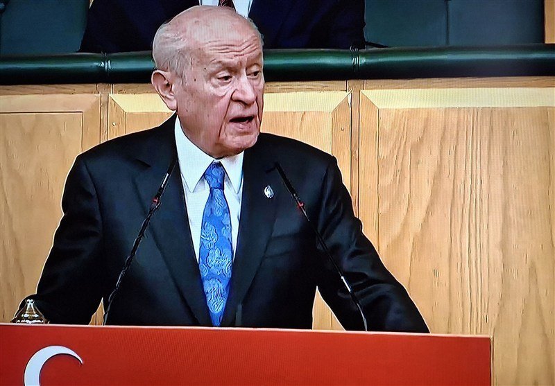 An elderly man in a dark suit and blue tie speaks at a podium displaying a partial white crescent on red, beneath a Turkish flag.