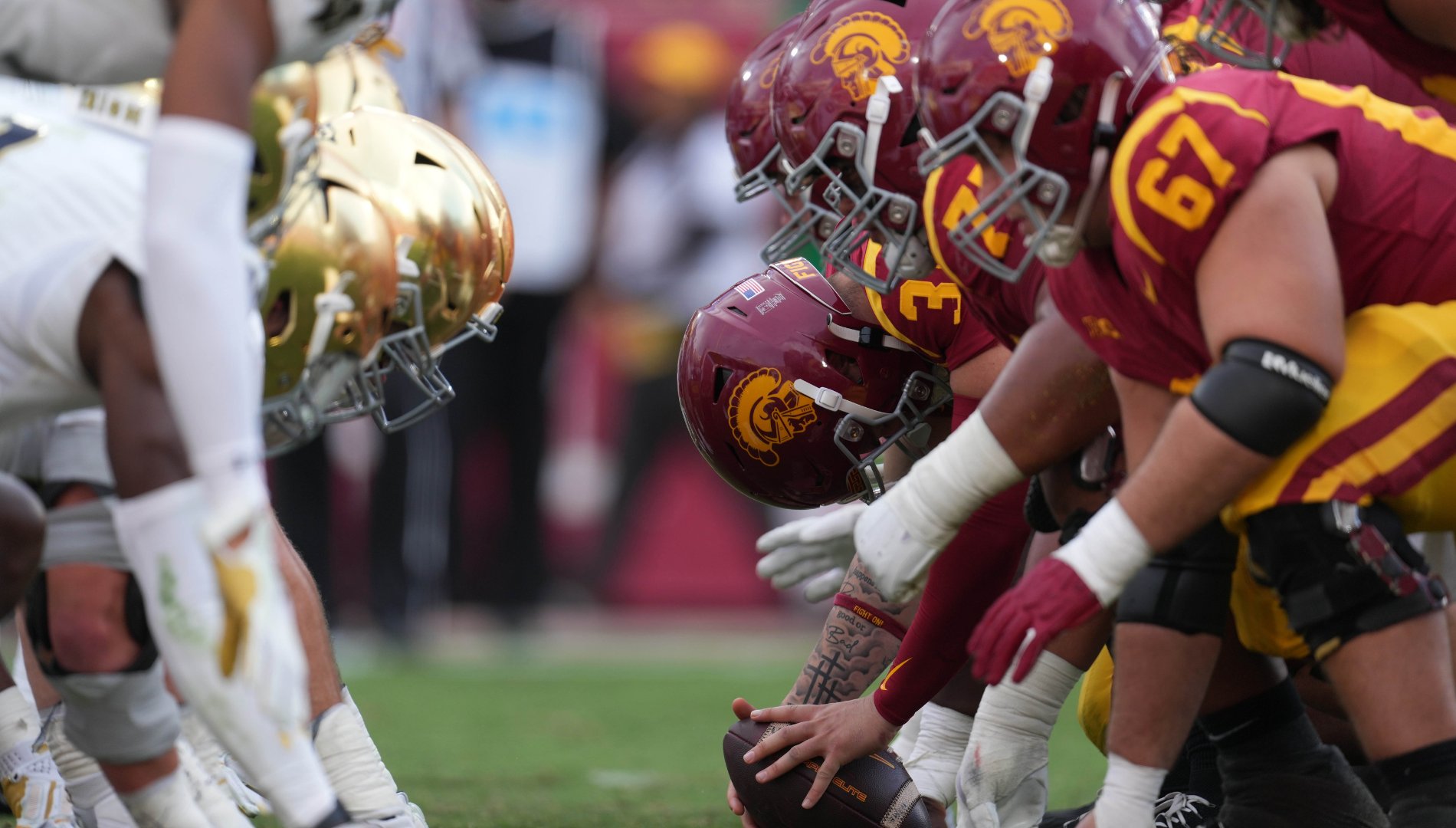 Football players in cardinal and gold helmets line up in a three-point stance against opponents wearing white and gold on the field.