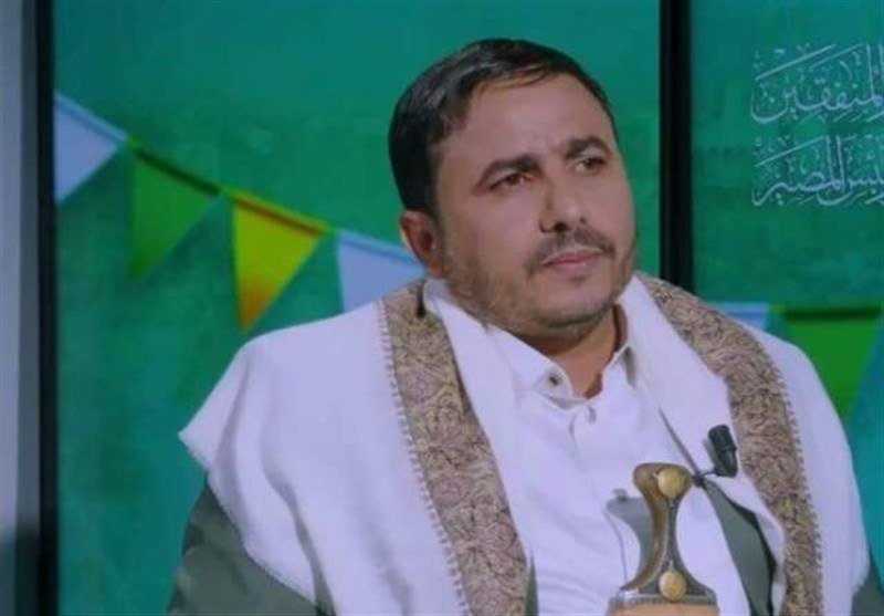 A man wearing a white shirt and embroidered shawl sits against a green backdrop with colorful triangular bunting, with a microphone visible nearby.