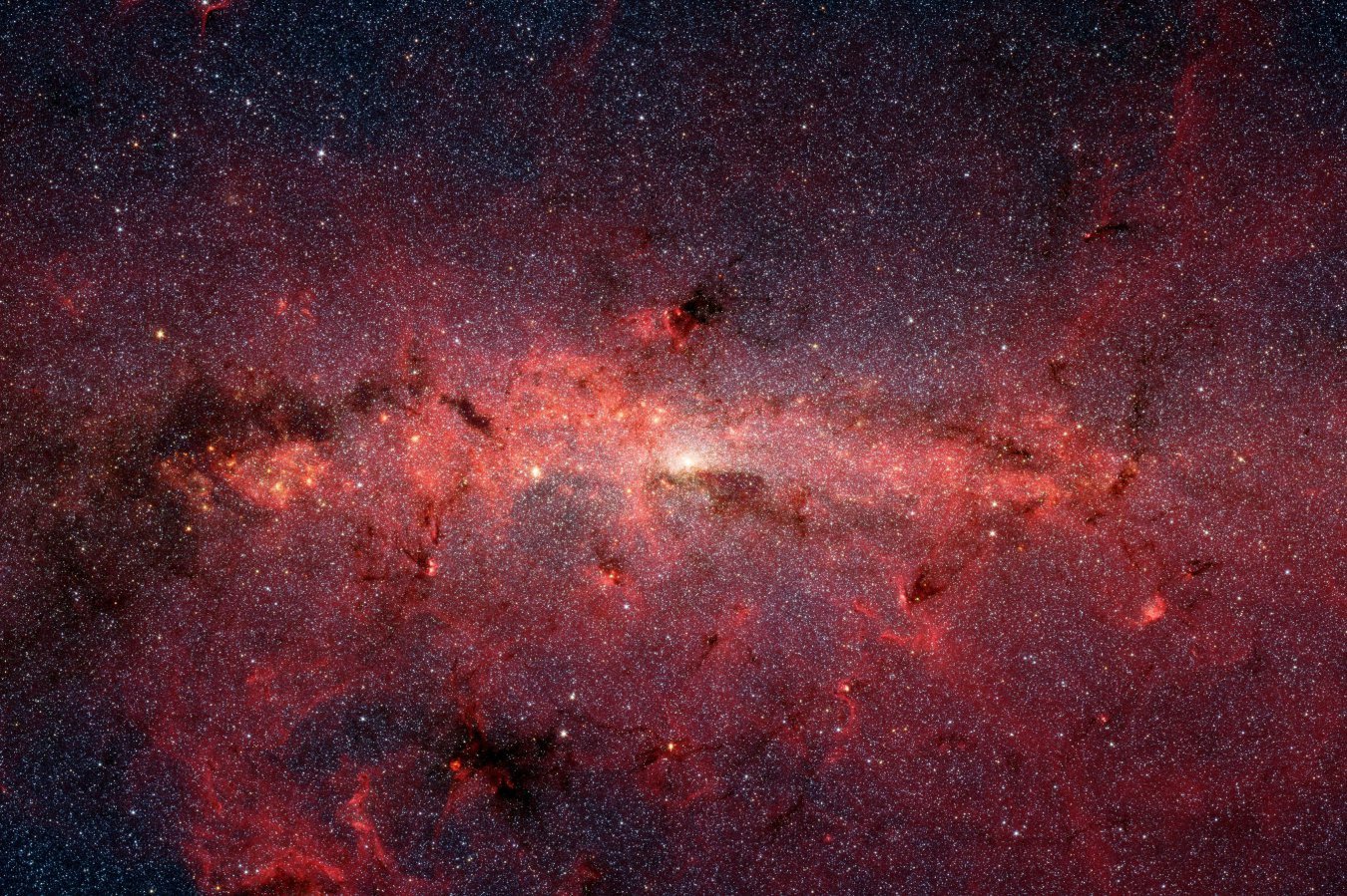 A glowing pink and red nebula with a bright central core stretches across a dark, star-filled sky, surrounded by dark dust clouds and countless distant stars.