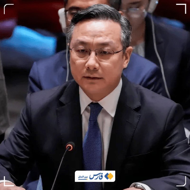 A bespectacled man in a dark suit and blue tie speaks into a microphone at a formal conference setting, with other attendees visible behind him.