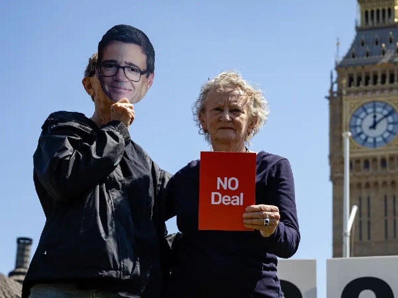 Two people protest outside a clock tower resembling Big Ben, one holding a large cutout mask of a man's face while the other holds a red "NO Deal" sign.