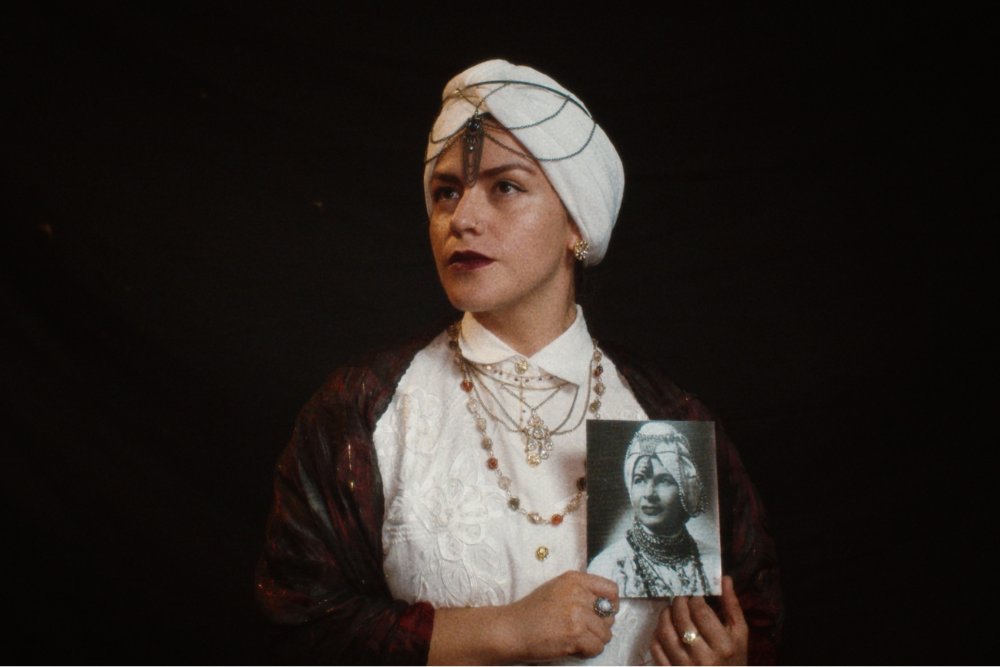 A person wearing a white turban, layered necklaces, and a dark shawl holds up a black-and-white portrait photograph of a similarly dressed figure against a black background.
