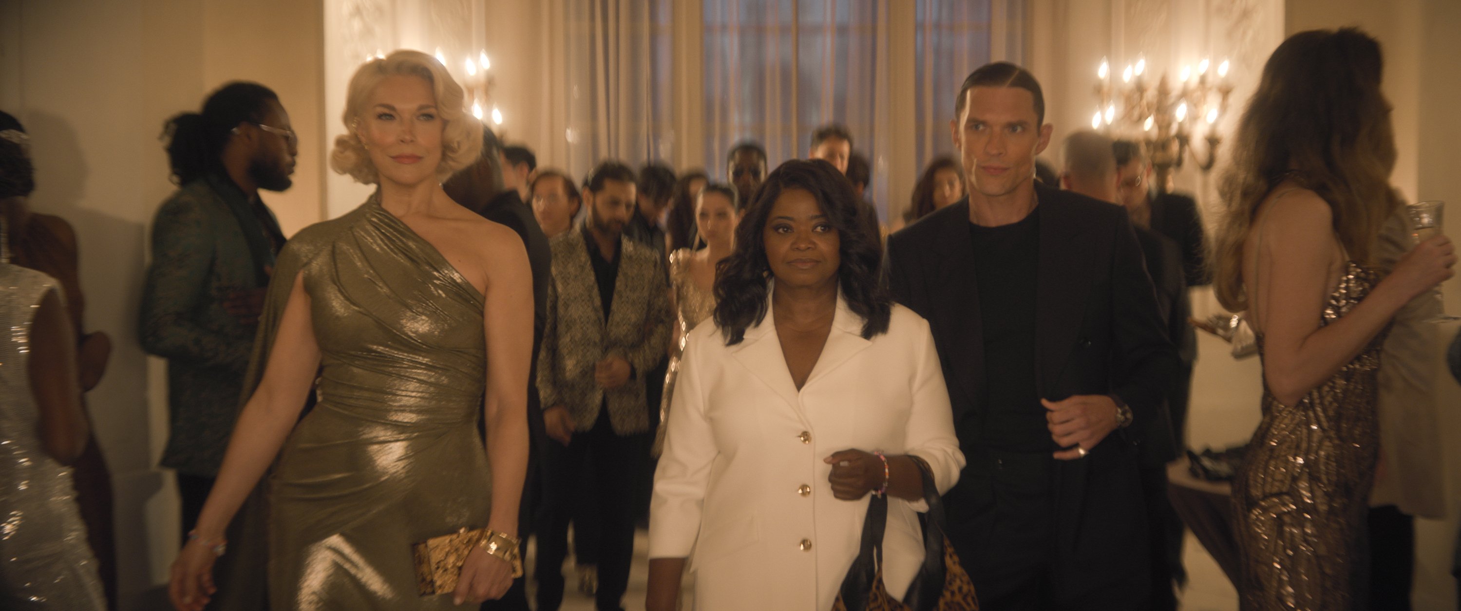 Three formally dressed individuals walk through a candlelit, chandelier-lit ballroom filled with other guests in evening wear.