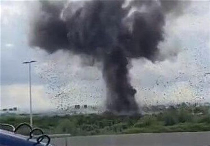 A large plume of dark smoke billows from an explosion in a grassy area, viewed through a rain-speckled window near a roadside with a streetlight.