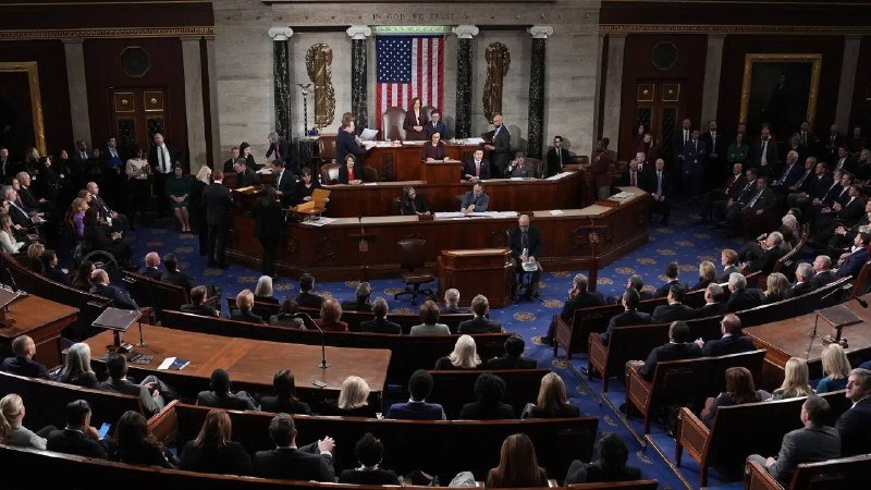 Officials gather in a formal legislative chamber with an American flag at the front, where individuals are speaking at a podium and presiding from an elevated bench.