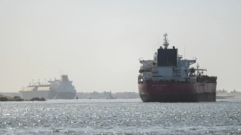 A large cargo ship sails on open water in the foreground with another tanker visible in the distance.