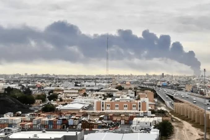 Plumes of dark smoke rise over a densely packed urban area with low-rise buildings, shipping containers, and a multi-lane highway under an overcast sky.