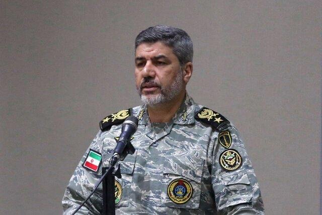 A man in a camouflage military uniform with insignia and shoulder boards speaks at a microphone against a plain background.