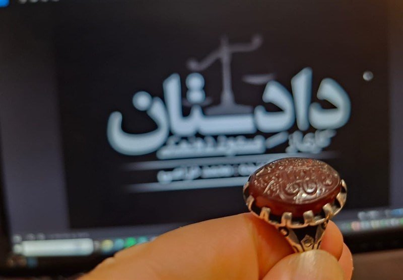 A hand holds up a ring with an ornate, engraved reddish-brown stone set in a silver-toned band, displayed against a blurred computer screen showing Persian text.