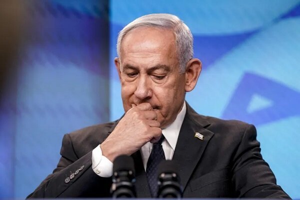 An older man with white hair, wearing a dark suit and blue tie, sits at a podium with microphones against a blue Star of David backdrop, his hand near his mouth.