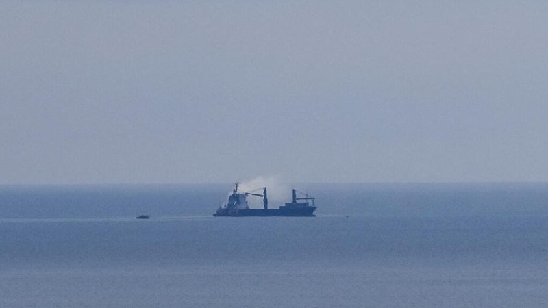 A large cargo ship with visible cranes sits on calm, hazy open water, with a smaller boat nearby.