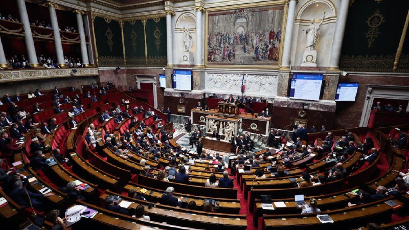 Lawmakers in formal attire fill the curved, semicircular seating of an ornate legislative chamber with classical statuary, columns, and a large mural above a central podium.
