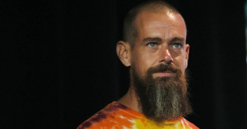 A bald, bearded man wearing an orange and yellow tie-dye shirt looks off to the side against a dark background.