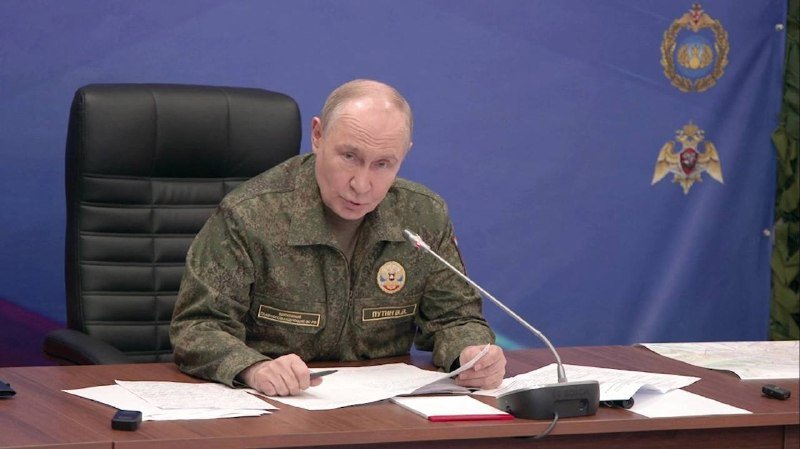 A man in a camouflage military uniform sits at a desk speaking into a microphone, with papers in front of him and a blue backdrop bearing emblems behind him.