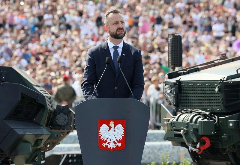 A bearded man in a dark suit speaks at a podium bearing a white eagle emblem, with military vehicles and a large crowd visible behind him.
