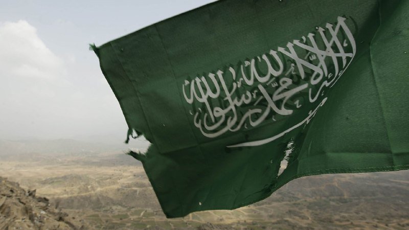 A green flag with white Arabic script and a sword emblem waves against a backdrop of rocky, mountainous terrain under a hazy sky.