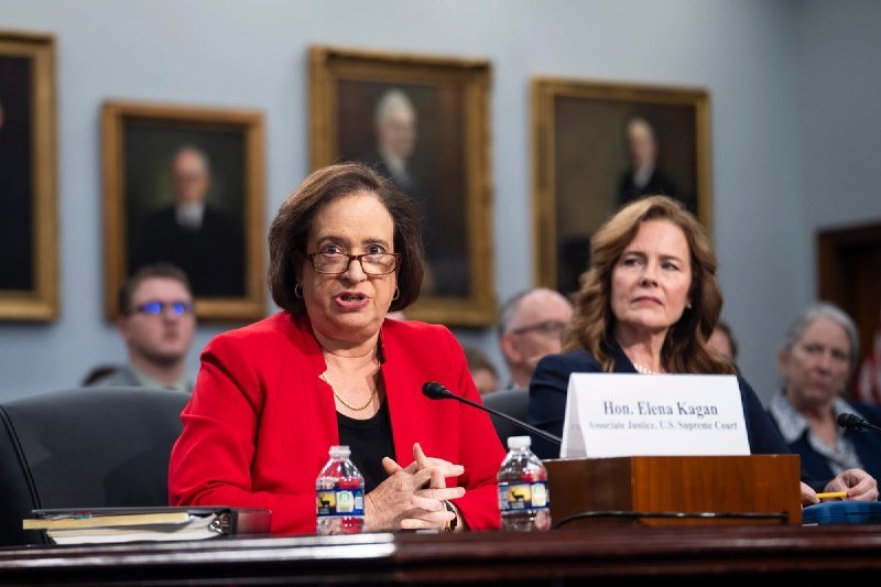 A woman in a red blazer speaks into a microphone at a witness table with a placard reading "Hon. Elena Kagan, Associate Justice, U.S. Supreme Court," seated beside another woman.