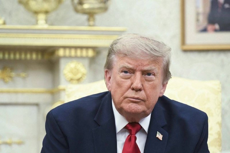 A gray-haired man in a dark suit and red tie sits in an ornate room with a gilded mantelpiece and framed artwork behind him.