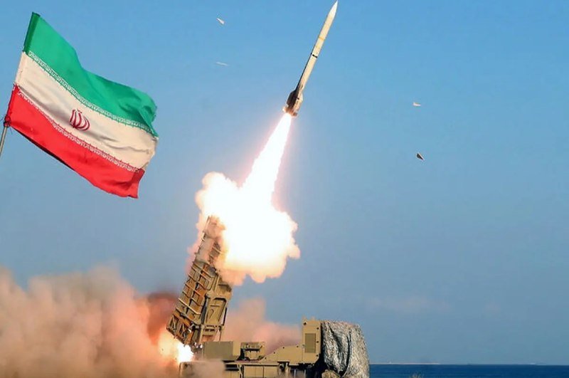A missile launches from a mobile launcher with a bright flame and smoke plume, while a green, white, and red flag waves in the foreground against a clear sky.