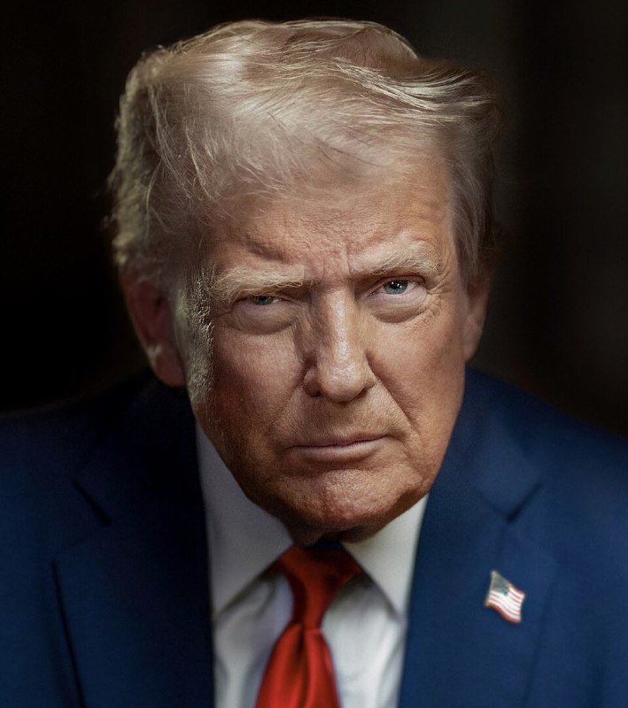 A man with blond hair wearing a navy blue suit, red tie, and an American flag pin looks directly at the camera against a dark background.