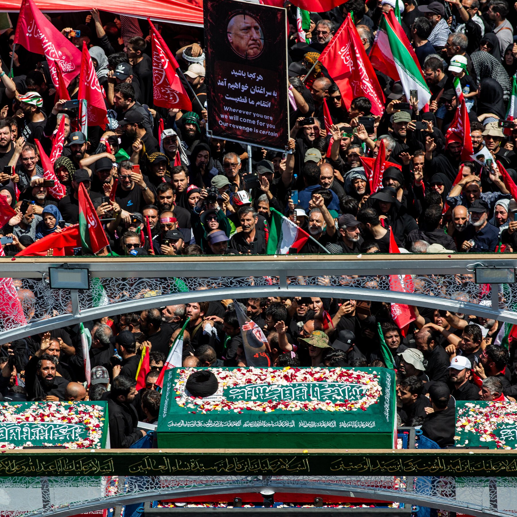 A massive crowd holds red flags and a portrait-bearing placard reading "Wherever you are we will come for you Our revenge is certain" around a flower-draped green coffin marked with Arabic script.