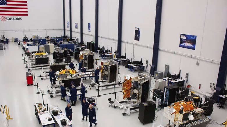 L3Harris logo and American flag displayed over an aerial view of a cleanroom facility where technicians assemble and inspect multiple spacecraft.