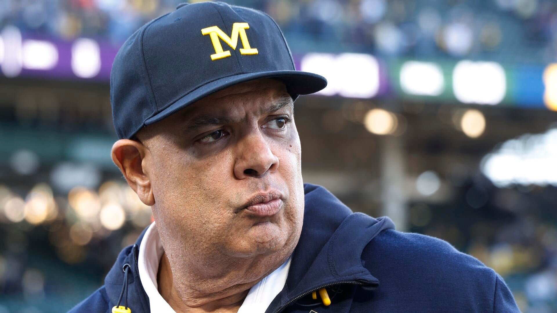 A man wearing a navy blue cap with a yellow "M" and matching hoodie looks off to the side with a serious expression in a blurred stadium setting.