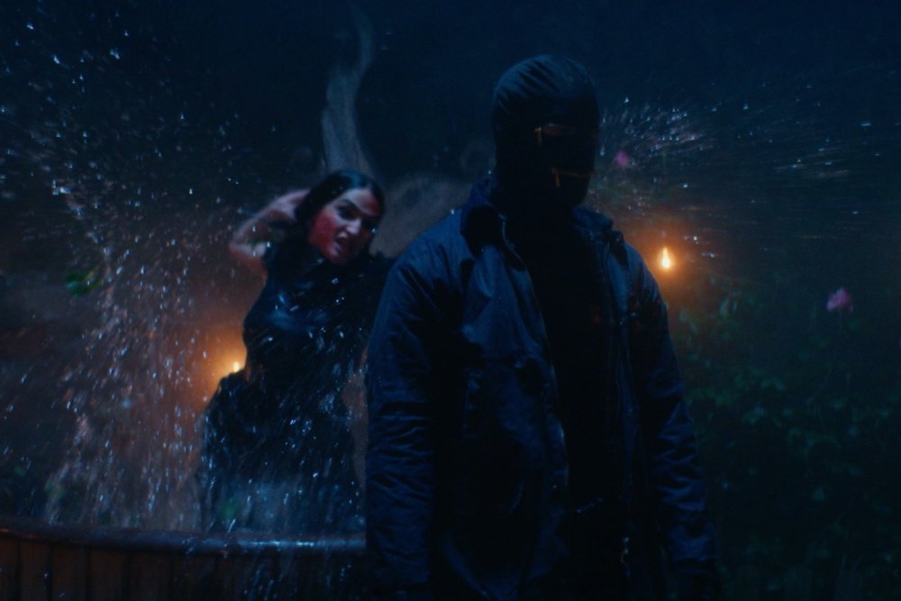 Two masked figures in dark jackets stand near a splashing water feature at night, illuminated by dim, warm background lighting.