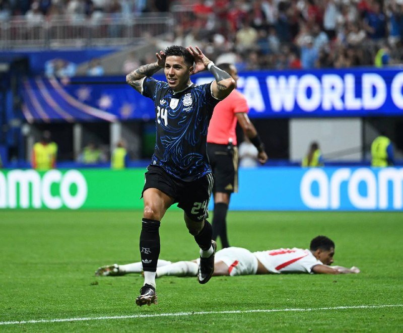 A soccer player wearing a dark blue #24 jersey cups his hands behind his ears in celebration, while an opponent lies face-down on the grass behind him.