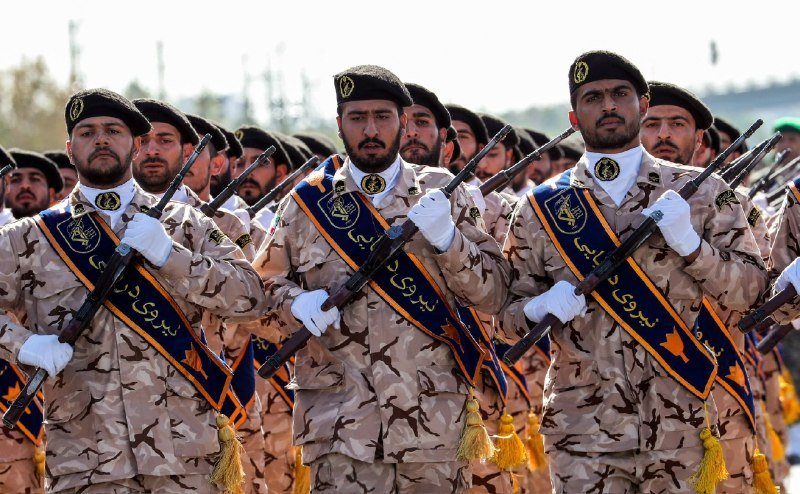 Soldiers in desert camouflage uniforms and black berets march in formation while carrying rifles, wearing blue sashes with Persian script.
