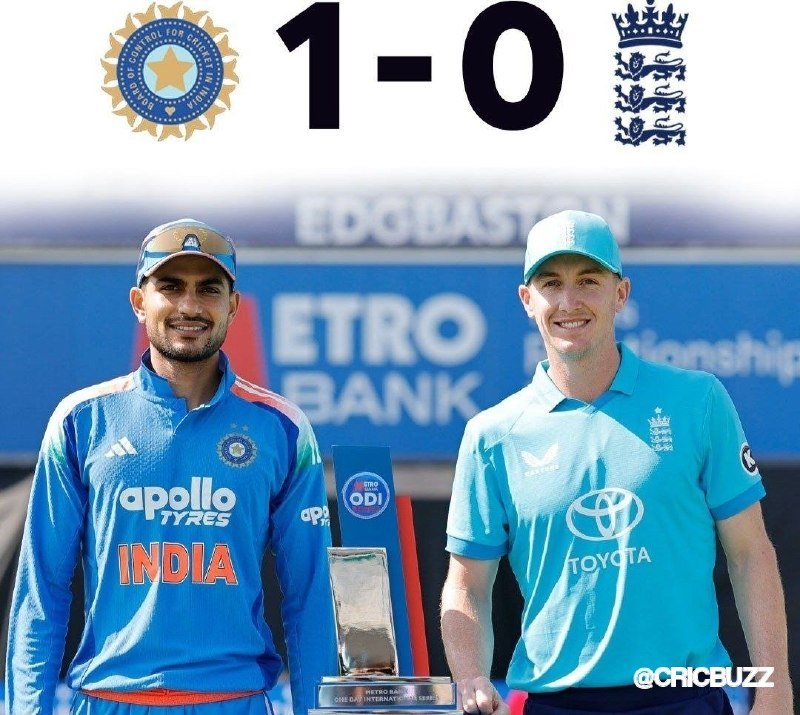 Two cricket captains in blue India and England jerseys pose with a trophy beside a "1-0" graphic displaying both team logos.