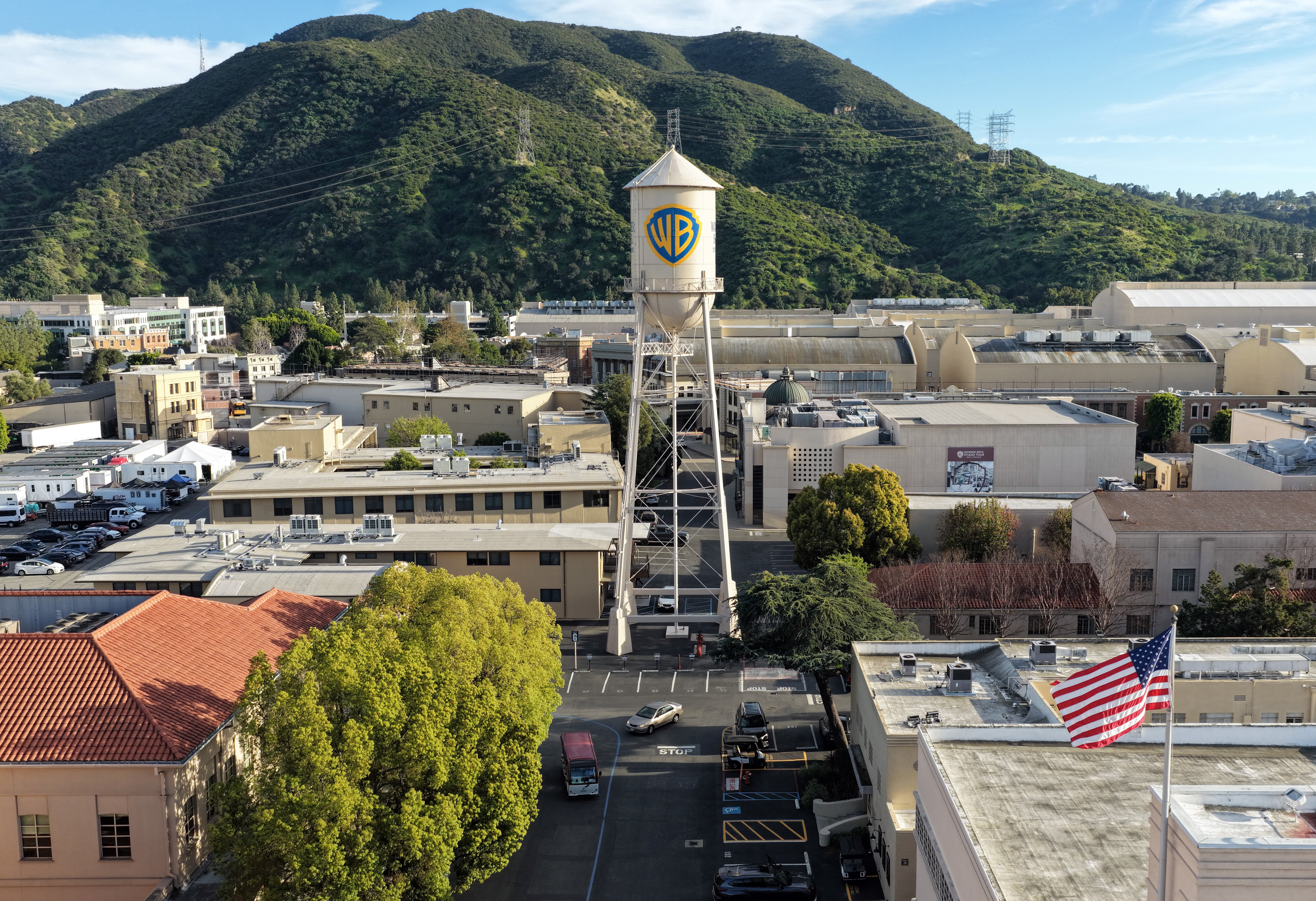 A WB-branded water tower stands above a studio lot's production buildings and parking lot, with forested mountains rising in the background.