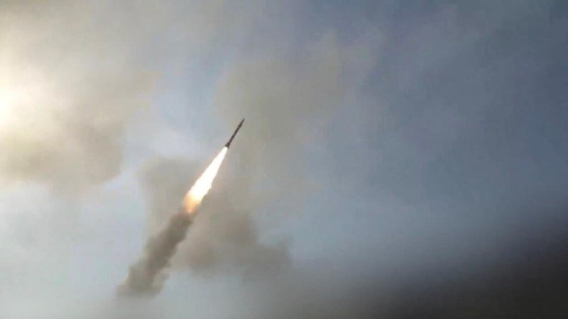 A missile ascends through clouds, leaving a bright flame and trailing smoke against a hazy sky.