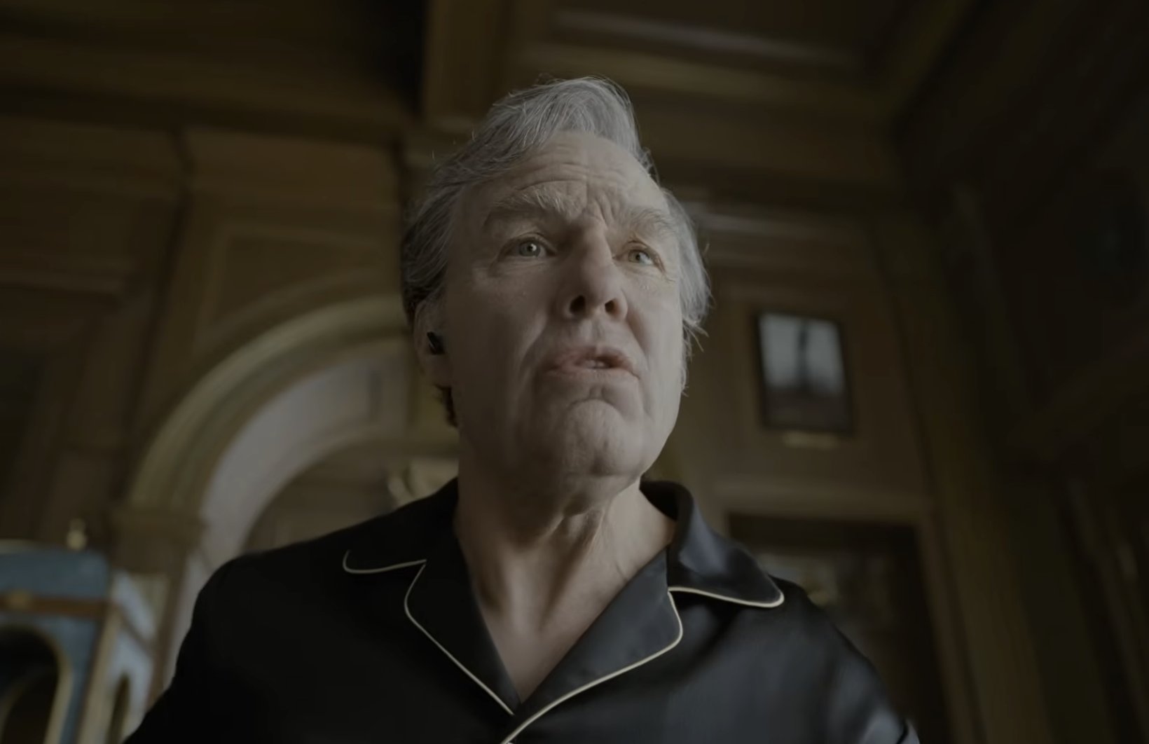 An older man with gray hair wears a dark, white-piped pajama-style shirt and a small earpiece, looking upward with pursed lips in a dimly lit wood-paneled room.