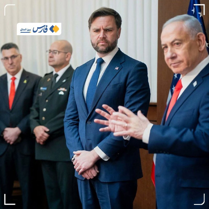Four men in formal attire stand in a room; one in a blue suit and tie stands center, while another gestures with his hands.