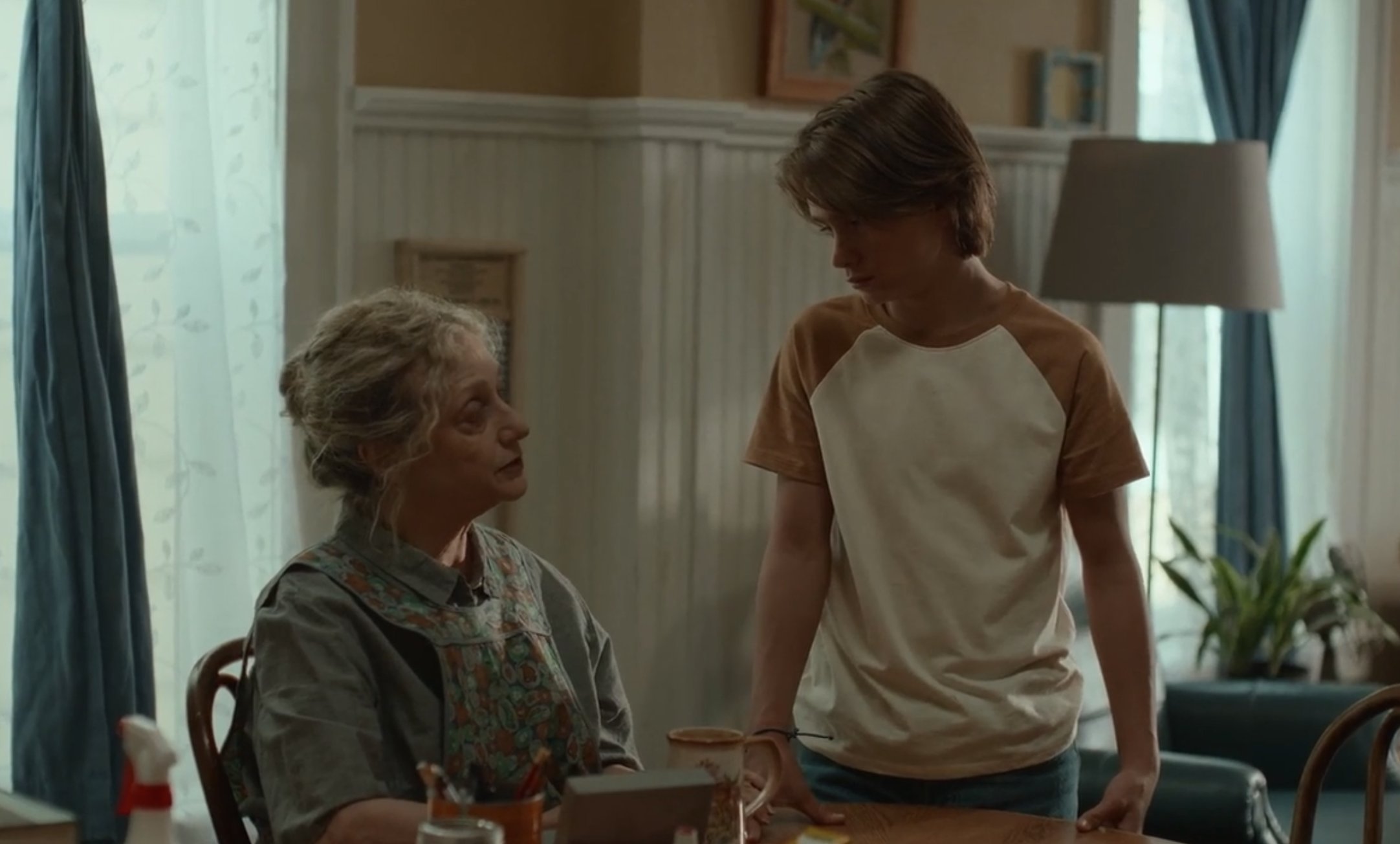 A seated older woman with curly hair looks up at a standing teenage boy across a dining table in a softly lit room with sheer curtains and a floor lamp.