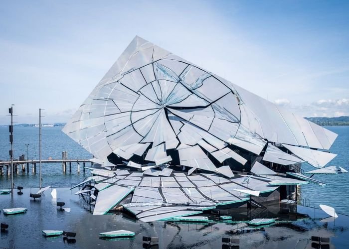 A large shattered crystal-like sculpture sits on a paved waterfront plaza beside a body of water under a partly cloudy sky.