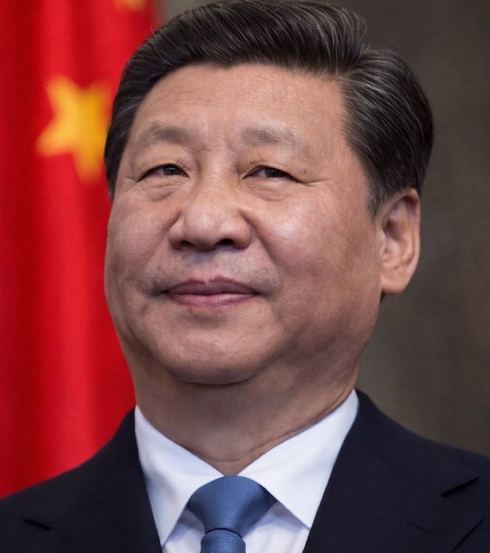 A man in a dark suit and light blue tie smiles in front of a red flag with yellow stars.