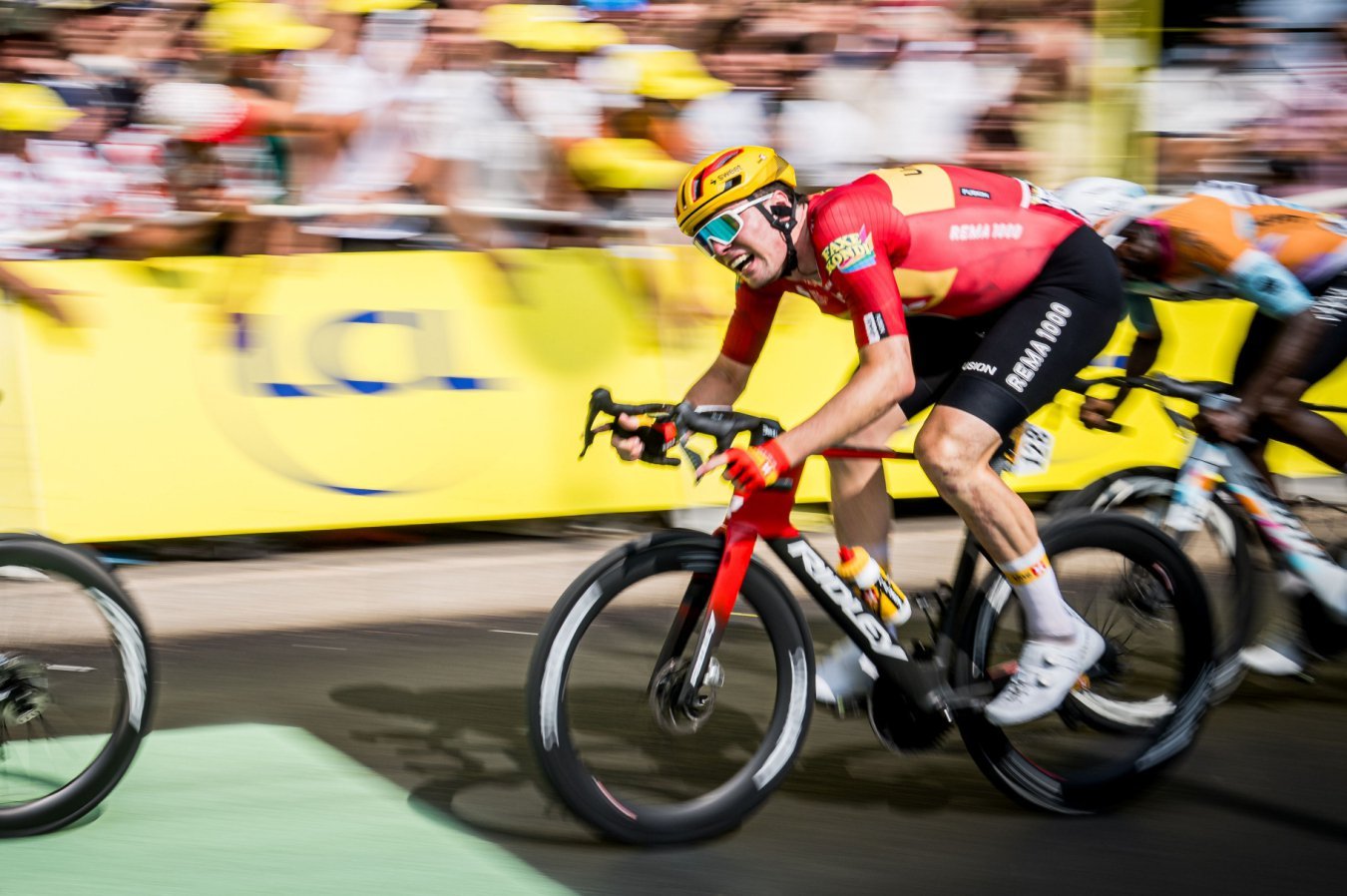 A cyclist in a red and yellow jersey sprints on a red bicycle past blurred spectators and yellow barriers, wearing a yellow helmet and reflective sunglasses.