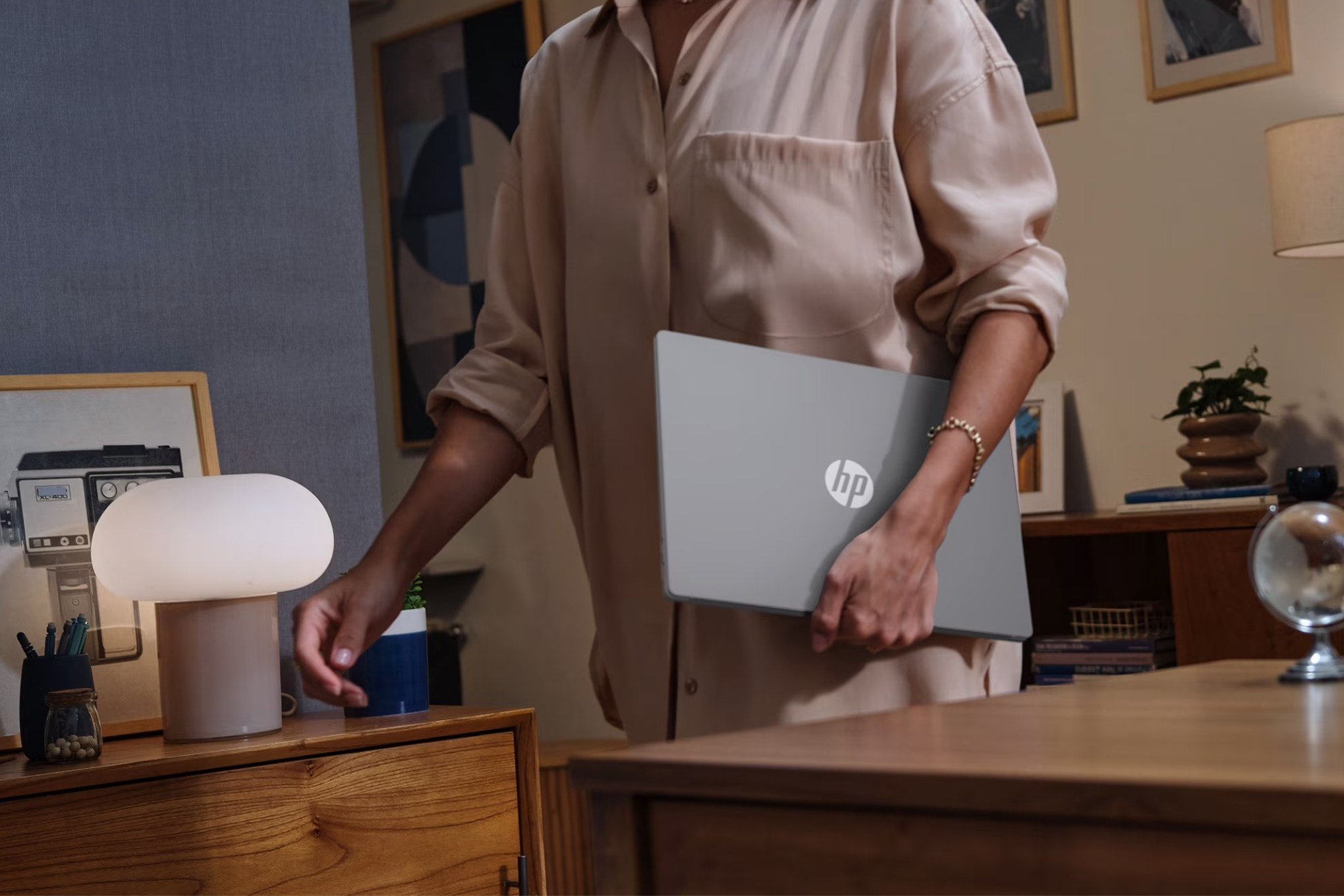 A person in a beige shirt carries an HP laptop while reaching toward a mushroom-shaped lamp on a wooden desk.